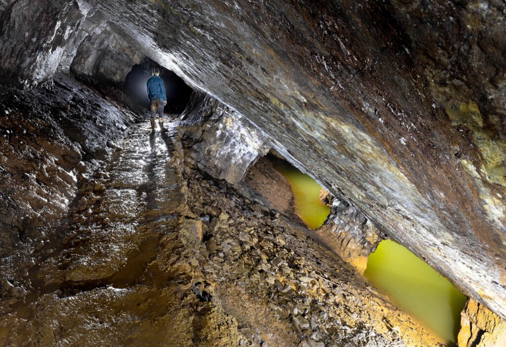 An explorer standing in a slanted mine tunnel beside pools of yellow water