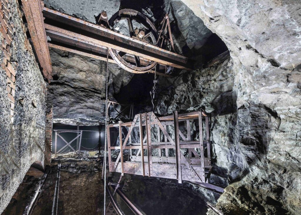 A rusty metal mine cage suspended from a pulley system inside a rocky underground shaft