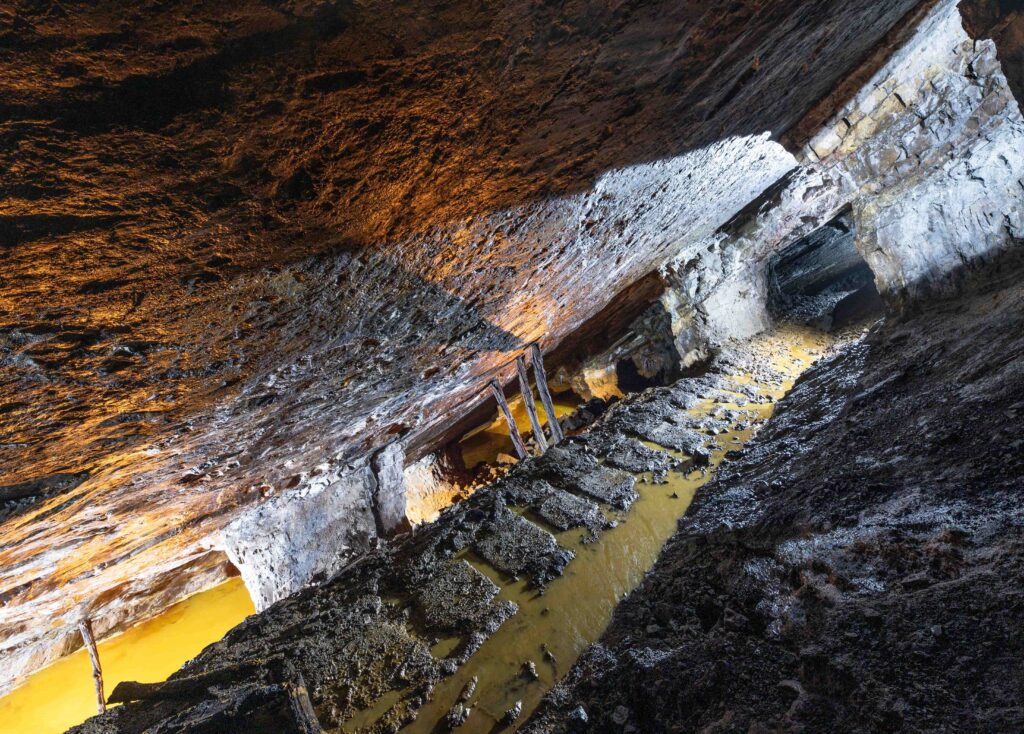 Galerie minière inclinée aux parois rocheuses sombres avec de l'eau jaunâtre au sol