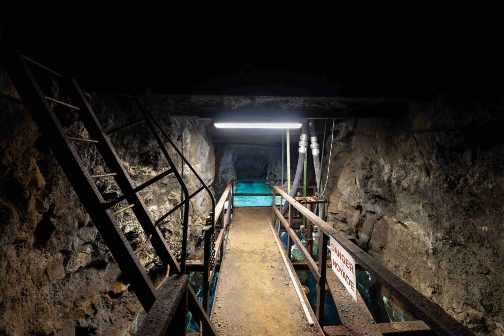 Illuminated metal footbridge over clear turquoise water inside a rocky mine tunnel