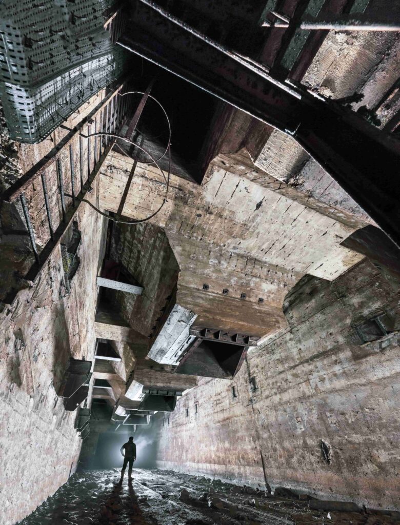 A person stands at the bottom of a mine shaft beneath concrete hoppers