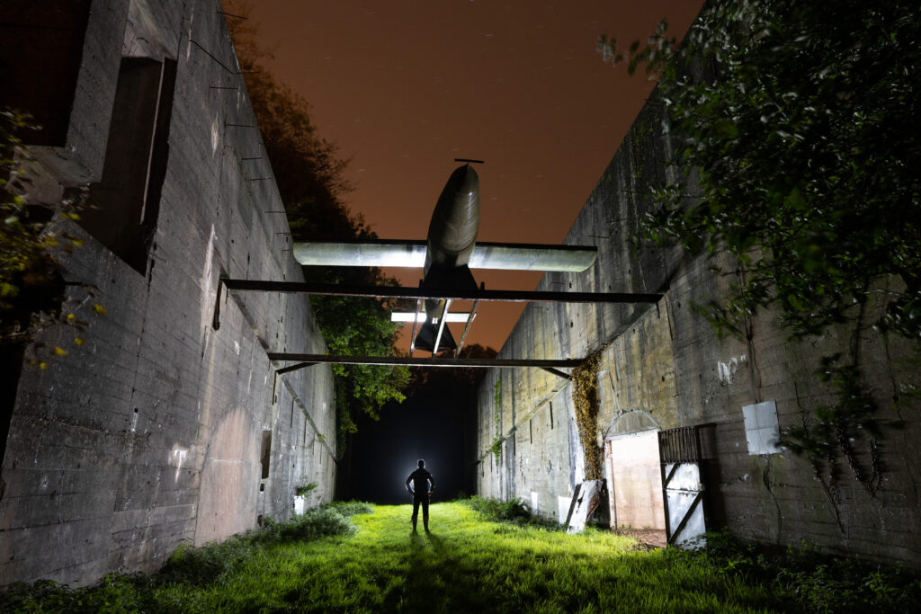 Un visiteur éclairé debout entre deux hauts murs en béton surmontés d'une réplique de V1