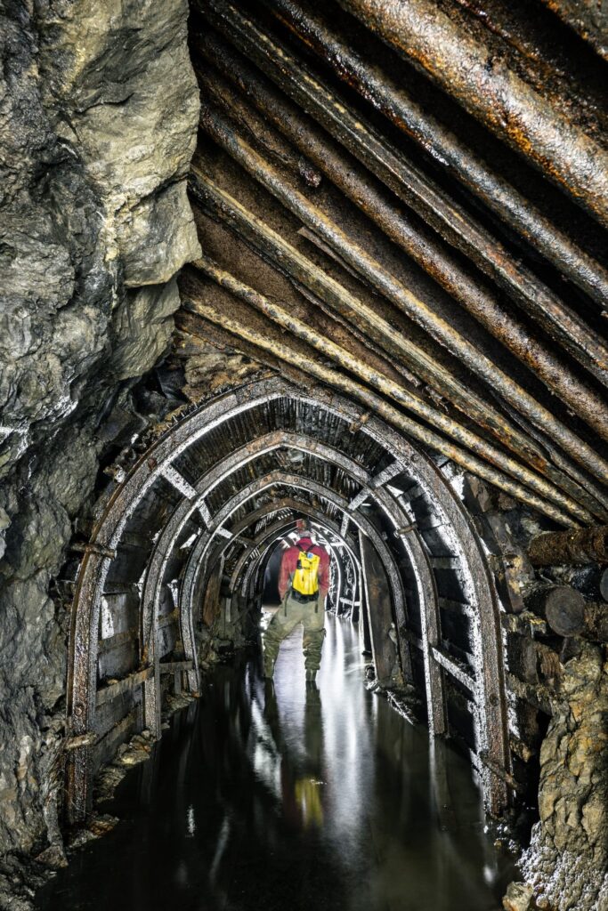 Personne équipée d'un casque et d'un gilet de sécurité marchant dans une galerie de mine étroite, soutenue par des arceaux métalliques, avec de l'eau au sol.